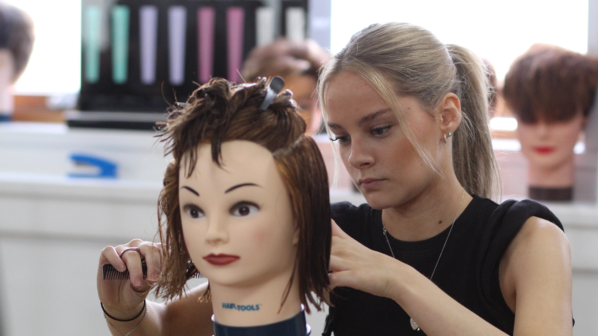Students practising hair cutting on mannequin heads in a classroom.