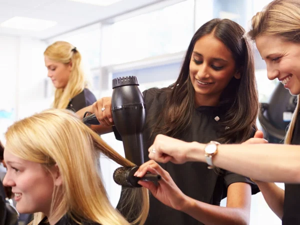 Learners practise hair styling in a training salon, using a hairdryer and brush while others observe nearby.