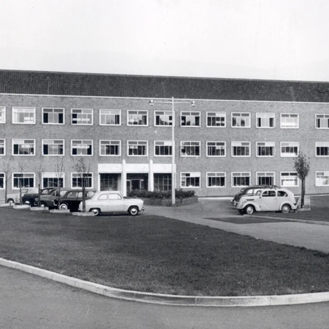 Black-and-white photograph of a large three-storey rectangular building with rows of windows, a central entrance, and several parked vintage cars in front.