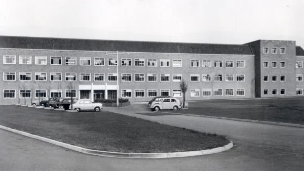Black-and-white photograph of a large three-storey rectangular building with rows of windows, a central entrance, and several parked vintage cars in front.