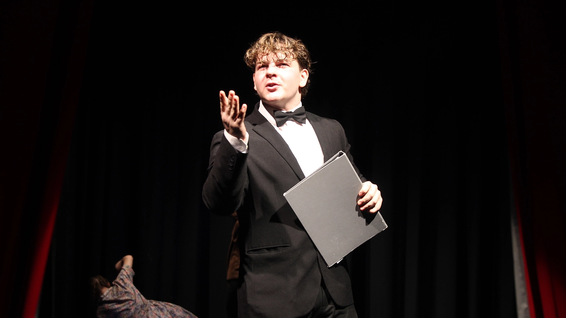 Performer in tuxedo gestures while holding a script folder under stage lighting.