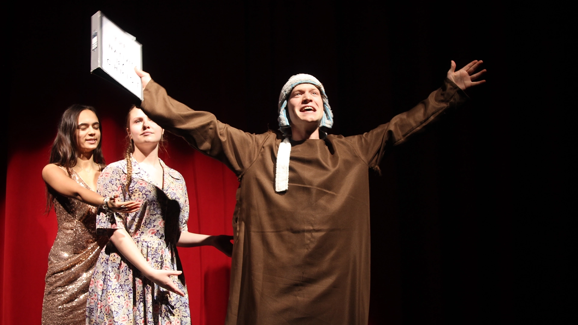 Performer in brown costume raises arms on stage while holding a sign, with two others presenting.
