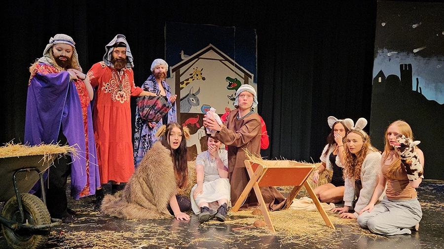 Cast in costume gather around a nativity scene with manger, hay and animal props on stage.