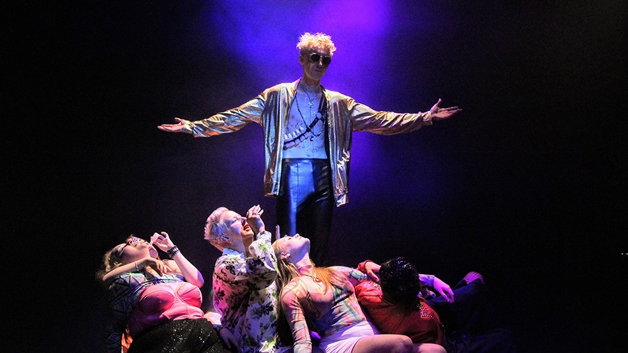 Performer in gold jacket stands centre stage with arms extended, others pose below.