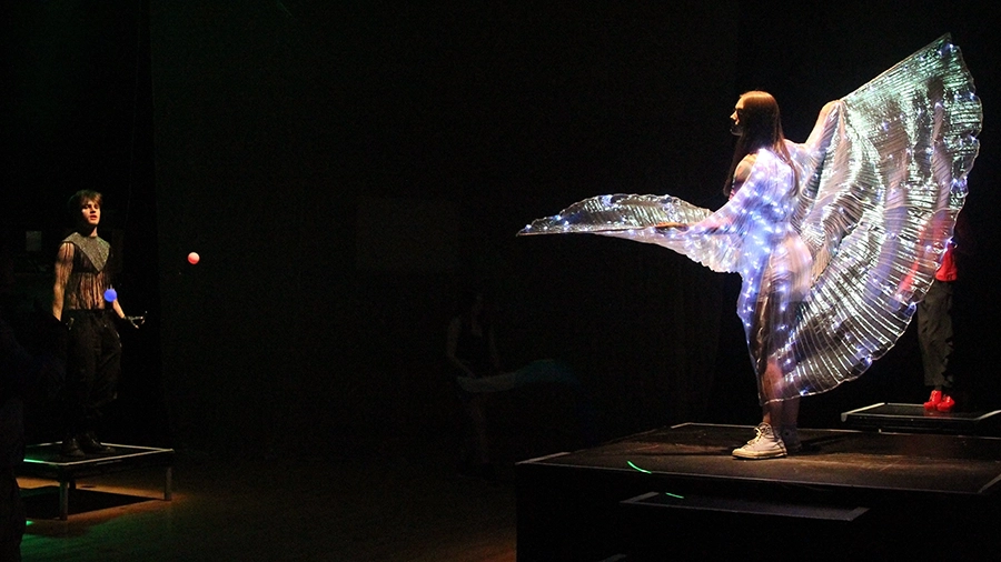 Performer wearing illuminated wings stands on platform while another juggles nearby.