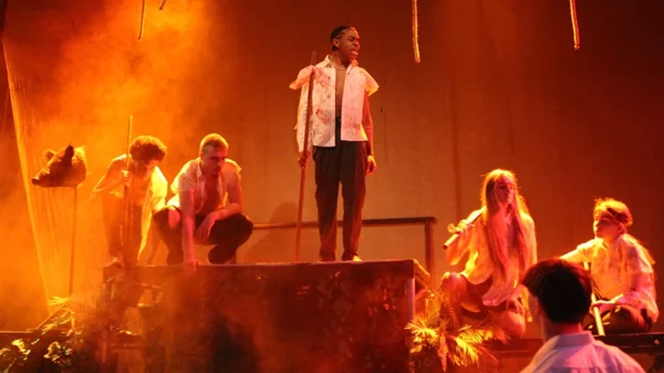 Group of performers stand on a platform under orange stage lighting with props and smoke effects.