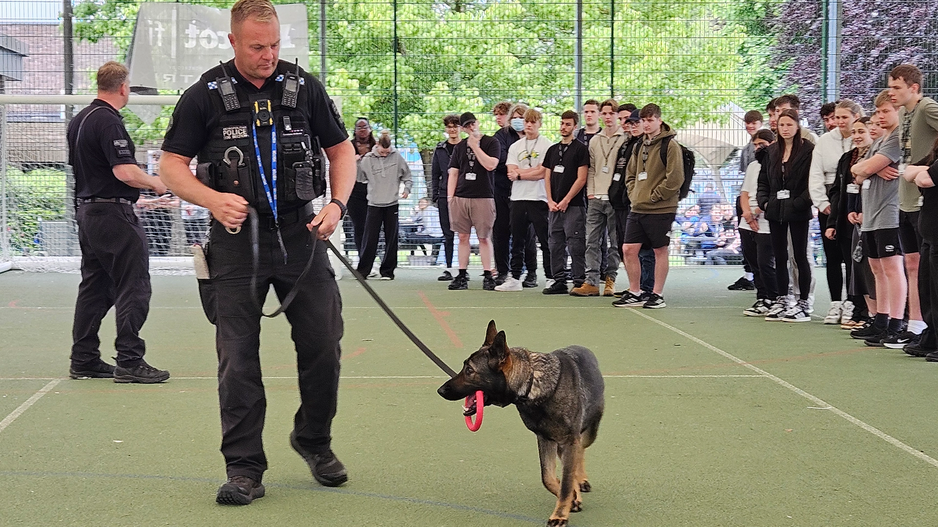 Police officer walking a dog on a lead while a group of students watch on a sports court.