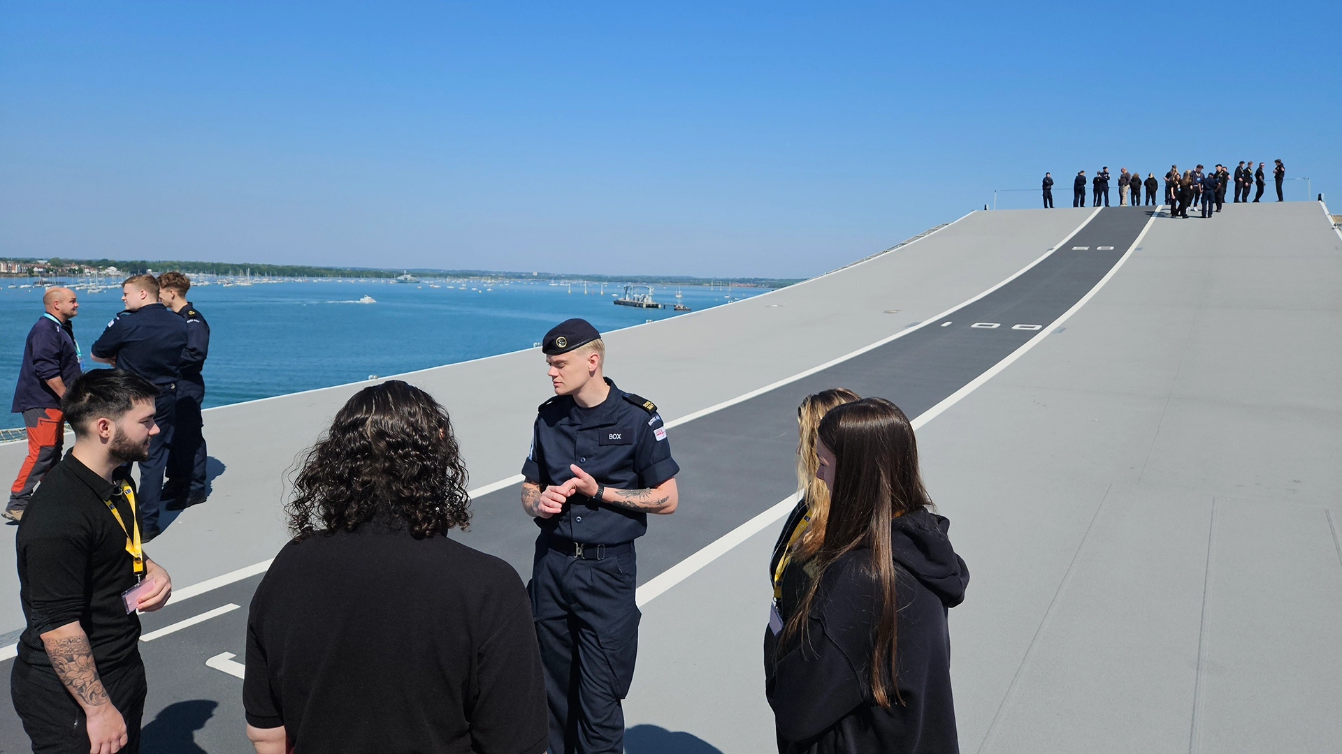 Uniformed personnel speaking with a small group on a large open deck overlooking the sea.