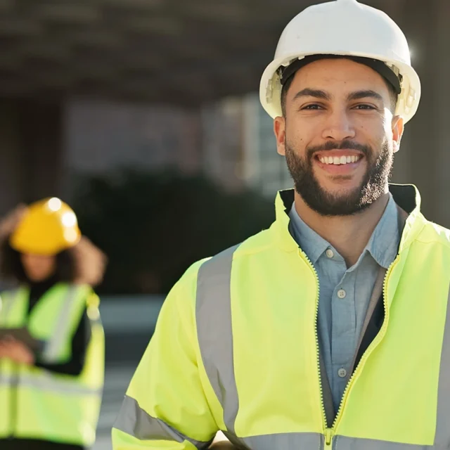 Person in a white hard hat and high-visibility jacket at a work site, with two others in safety gear in the background.