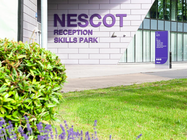 The exterior of the Nescot Skills Park building with large purple signage reading “NESCOT Reception Skills Park.” The scene includes landscaped greenery and modern angular architecture.