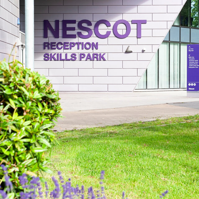 The exterior of the Nescot Skills Park building with large purple signage reading “NESCOT Reception Skills Park.” The scene includes landscaped greenery and modern angular architecture.