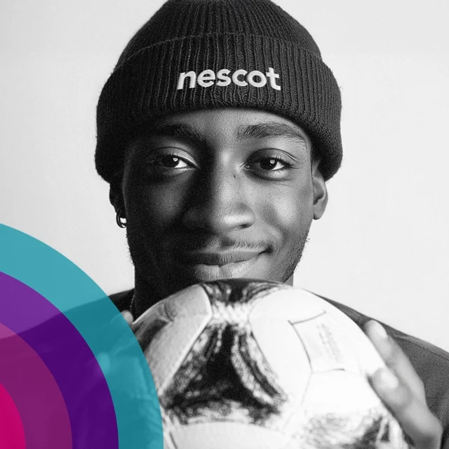 A person wearing a beanie with the word ‘nescot’ on it holds a football close to the camera while standing against a plain background.