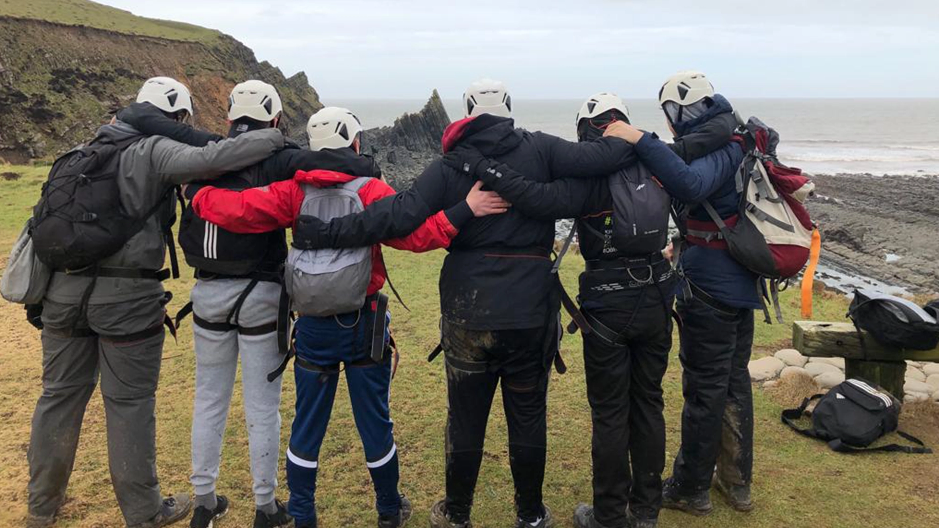 Group wearing helmets standing arm in arm on a coastal cliff path overlooking the sea.