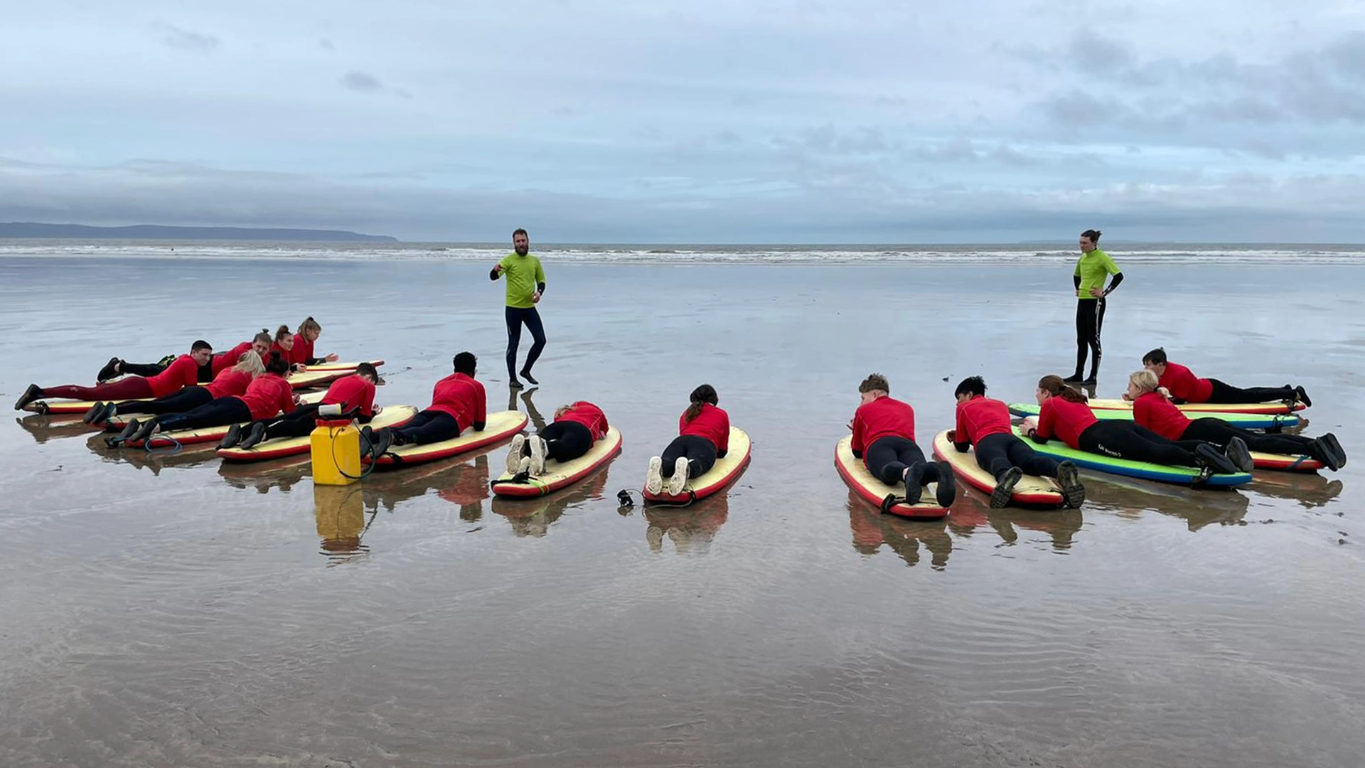 Group lying on surfboards on a beach while an instructor stands nearby.