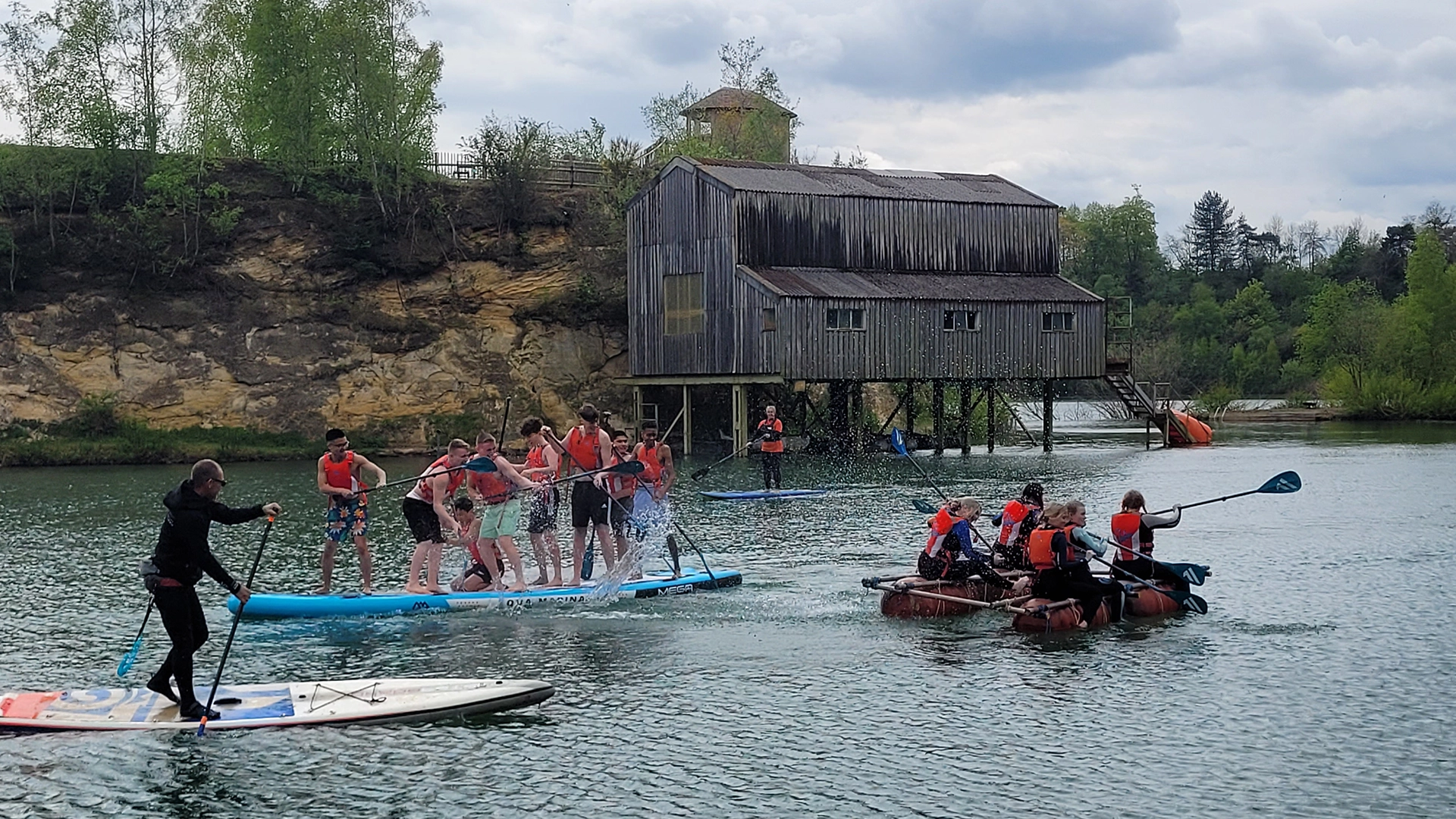 Group on paddleboards and a raft on a lake near a wooden building on stilts.