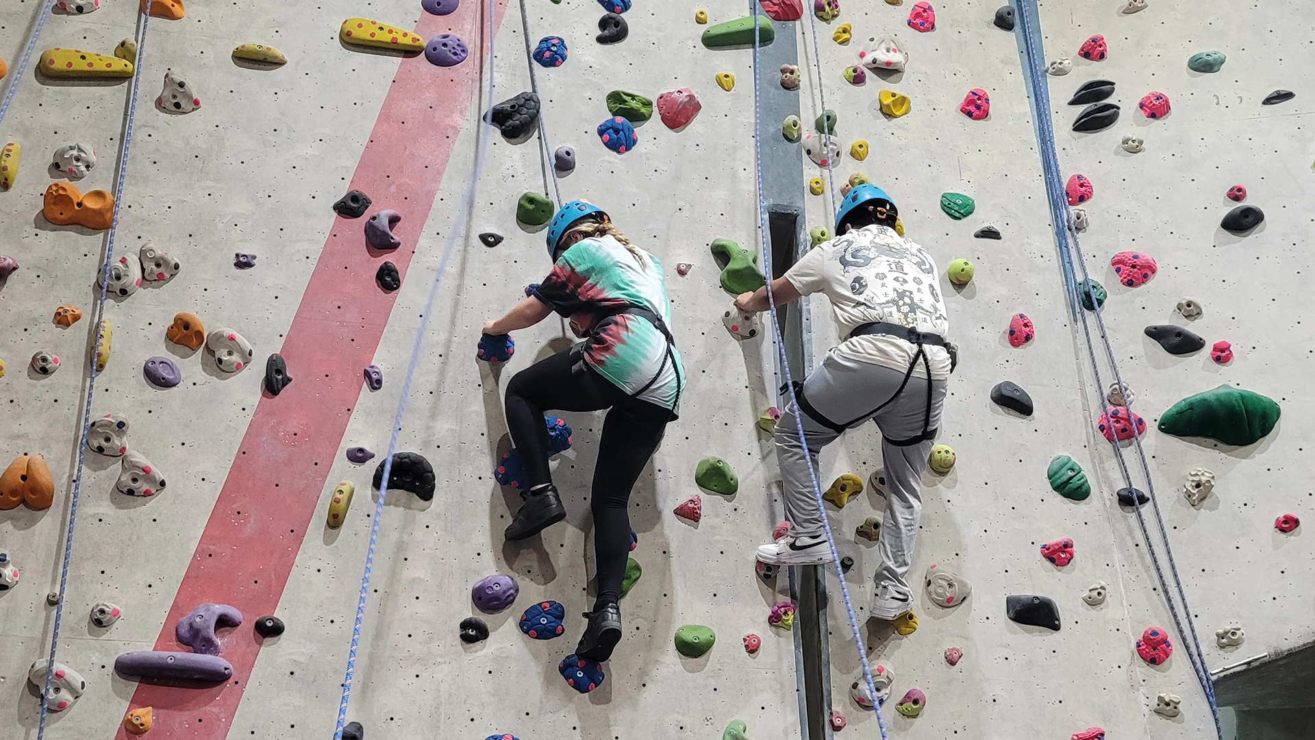 Two people climbing an indoor rock wall with safety ropes.
