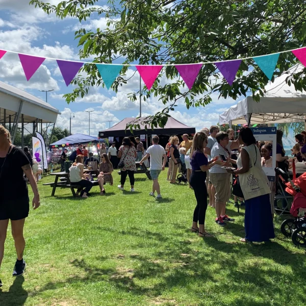 People attending an outdoor community fair with stalls, picnic benches, and bunting on a sunny day.