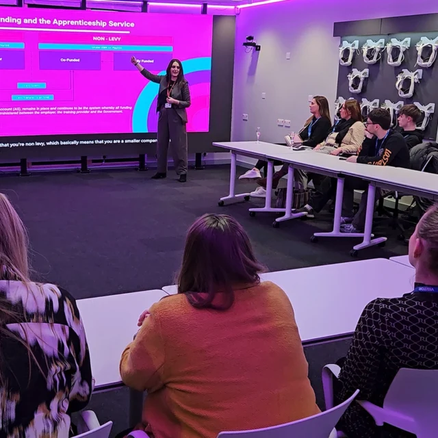 Presenter standing at the front of a classroom pointing to a large screen displaying apprenticeship funding information, while attendees sit around tables listening.