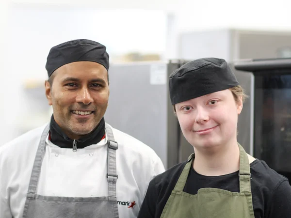 A ched and a trainee in a commercial kitchen wearing chef hats and aprons, standing side by side in front of catering equipment.