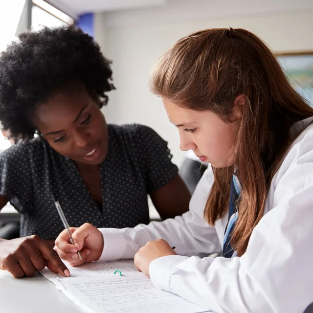 Teacher guiding a student writing in a workbook at a desk in a classroom with learning materials in the background.