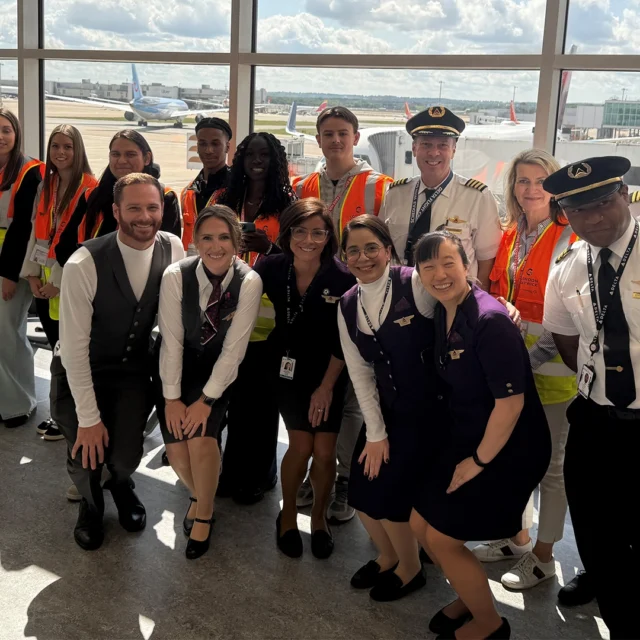 Group of students and aviation staff in high-visibility vests and airline uniforms posing in an airport terminal, with aircraft visible through large windows behind them.