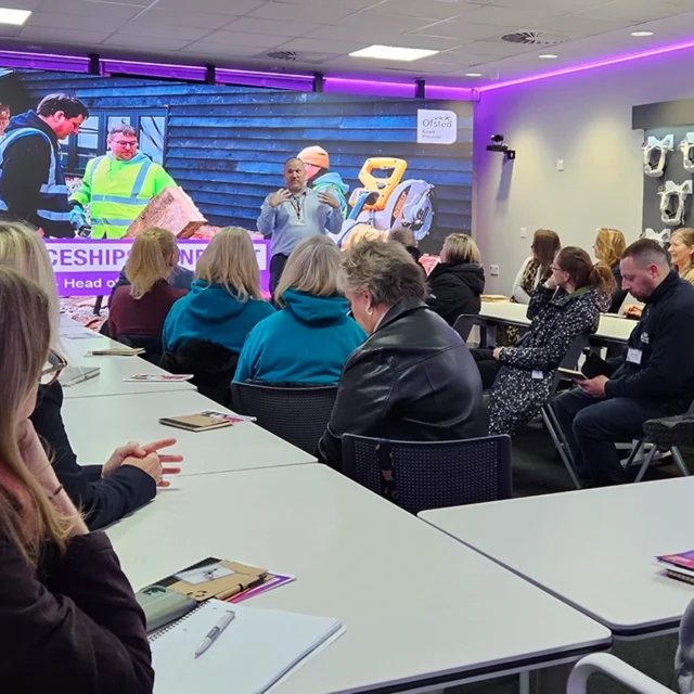 People seated at tables in a classroom watch a presenter speaking at the front, with a large screen displaying construction and apprenticeship imagery behind them.