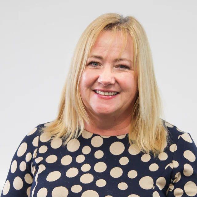 Portrait headshot of Yvonne Spencer a smiling woman with straight blonde hair, wearing a navy top with large cream polka dots, against a light grey background