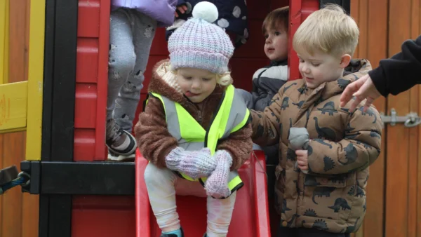 Children in outdoor play area, with toddlers in coats and hats taking turns on a red slide while supervised by an adult.