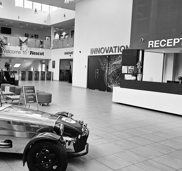 A modern Nescot atrium area with a reception desk, large windows, and a sports car parked beside visitor chairs.