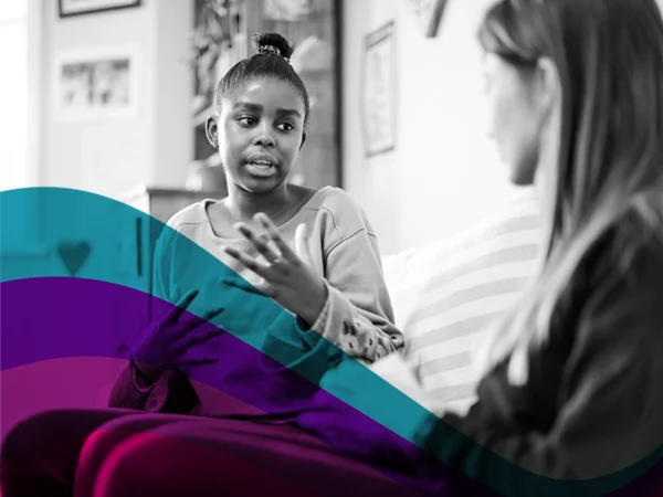 Black-and-white photo of a student sitting on a sofa, speaking and gesturing with their hands while another person listens, suggesting a one-to-one support conversation; teal and pink graphic overlay in foreground.