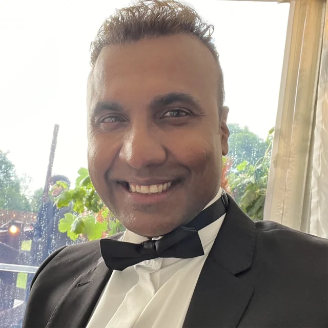Person in a black tuxedo and bow tie looking towards the camera indoors, with a window showing greenery in the background.