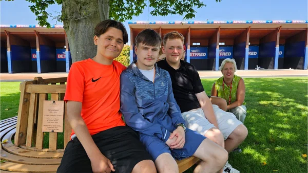 Three Nescot students sit on the circular wooden Pat Eddery memorial bench, while Nescot CEO and Principal Julie Kapsalis kneels alongside. The bench surrounds a large tree on the grassy area of the Parade Ring at Epsom Downs Racecourse.