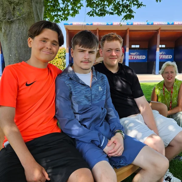 Three Nescot students sit on the circular wooden Pat Eddery memorial bench, while Nescot CEO and Principal Julie Kapsalis kneels alongside. The bench surrounds a large tree on the grassy area of the Parade Ring at Epsom Downs Racecourse.
