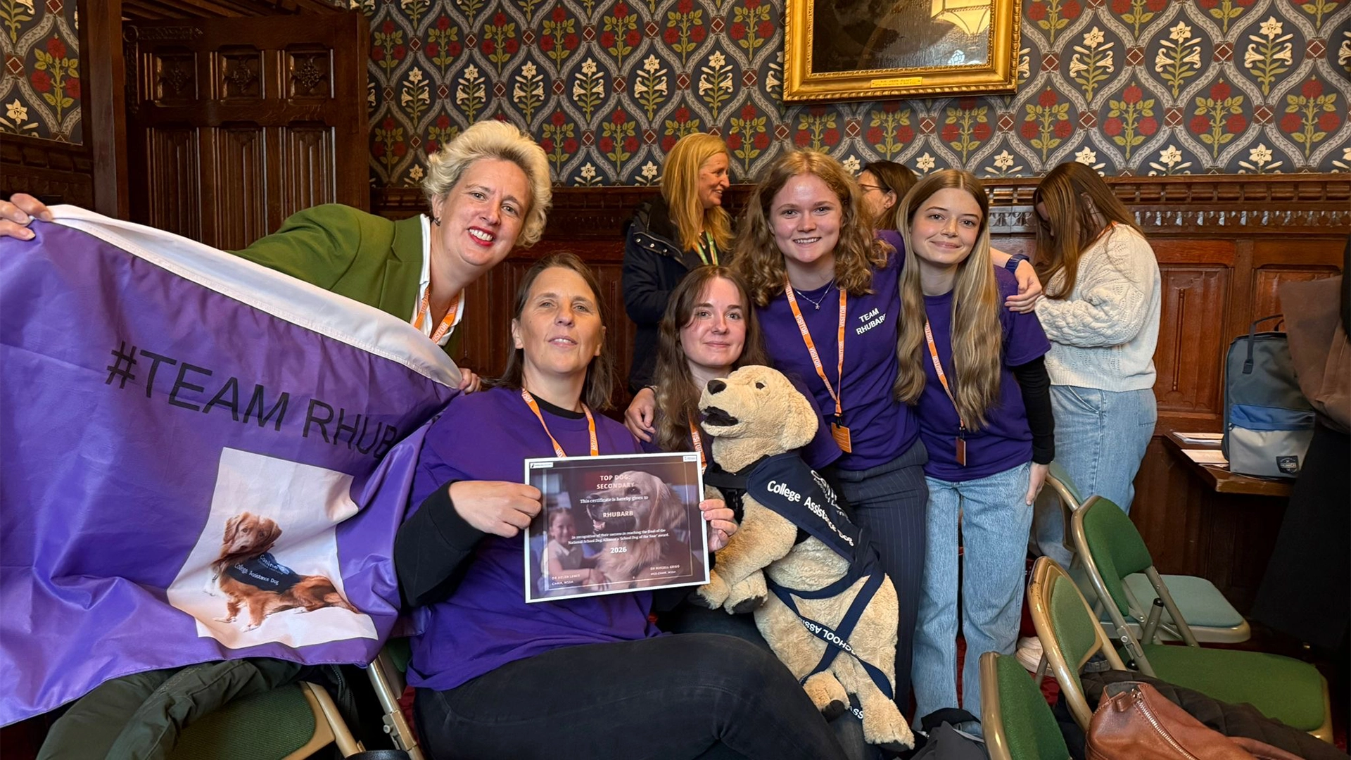 A group photo of Nescot staff and students the School Dog of the Year 2026 awards ceremony held at the UK House of Lords. They are holding a certificate and a purple "#Team Rhubarb" banner alongside a toy assistance dog wearing a college assistance dog jacket. The group is gathered in an ornate wood-panelled room with patterned wallpaper.