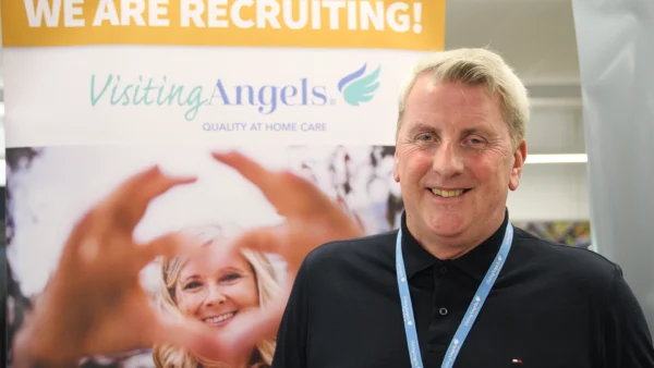 Chris James from Visiting Angels, wearing a black polo shirt and a blue lanyard stands in front of a Visiting Angels recruitment display. The banner behind them features the Visiting Angels logo, the text “WE ARE RECRUITING!” and “QUALITY AT HOME CARE,” along with an image of hands forming a heart shape around a smiling adult. The photo is taken indoors at a Nescot careers fair.