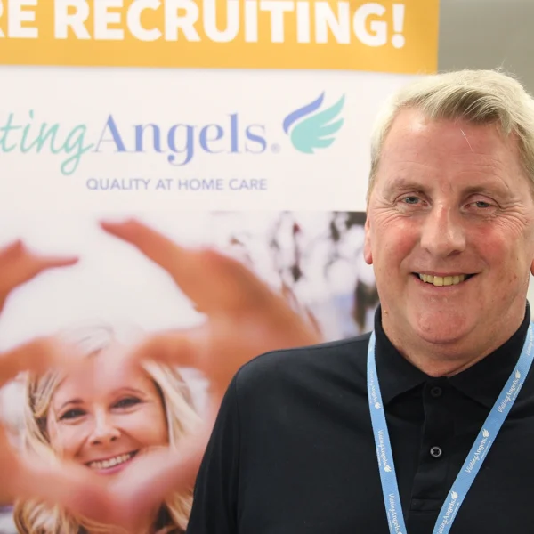 Chris James from Visiting Angels, wearing a black polo shirt and a blue lanyard stands in front of a Visiting Angels recruitment display. The banner behind them features the Visiting Angels logo, the text “WE ARE RECRUITING!” and “QUALITY AT HOME CARE,” along with an image of hands forming a heart shape around a smiling adult. The photo is taken indoors at a Nescot careers fair.