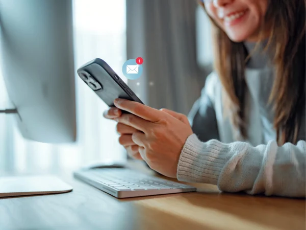A person sits at a desk holding a smartphone with both hands, looking at a screen that shows a new‑message notification. A computer monitor, keyboard, and mouse are on the desk, and soft daylight comes through a window in the background.