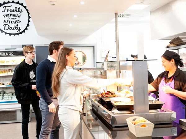 People queue at nescot refectory food counter while a staff member serves hot food from metal trays. A sign on the wall reads “Freshly prepared in our kitchen daily,” with refrigerated shelves and overhead lighting visible in the background.
