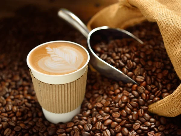 A takeaway cup of coffee with leaf-shaped latte art sits among scattered roasted coffee beans, next to a metal scoop and an open burlap sack filled with beans, under warm, soft lighting.
