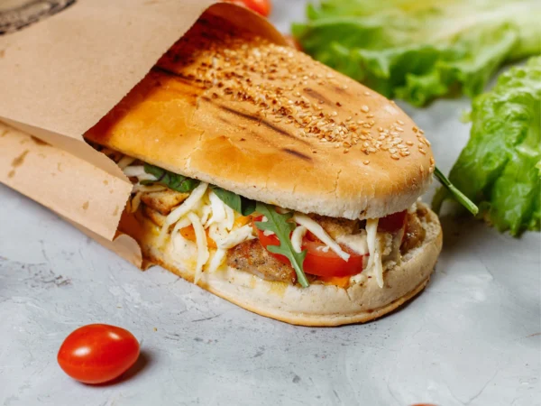 A toasted panini partially wrapped in brown paper, filled with crispy, cheese, sliced tomato, and rocket. The sandwich sits on a light stone surface with loose lettuce leaves and cherry tomatoes scattered around.