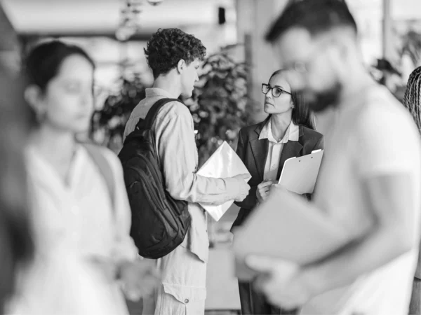 Student speaking with a member of staff while holding documents in a busy college environment, with other students in the background.