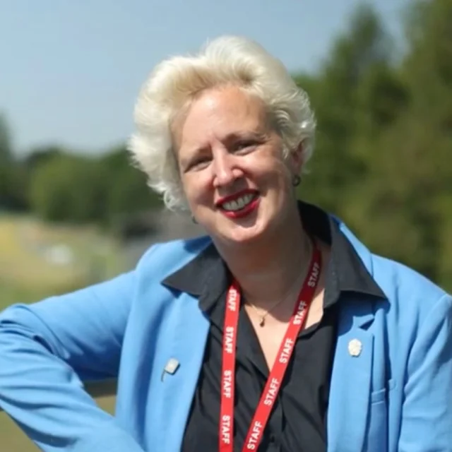 Principal Julie Kapsalis MBE wearing a blue blazer and red lanyard, leaning on a metal gate outdoors with a grassy field and trees in the background.