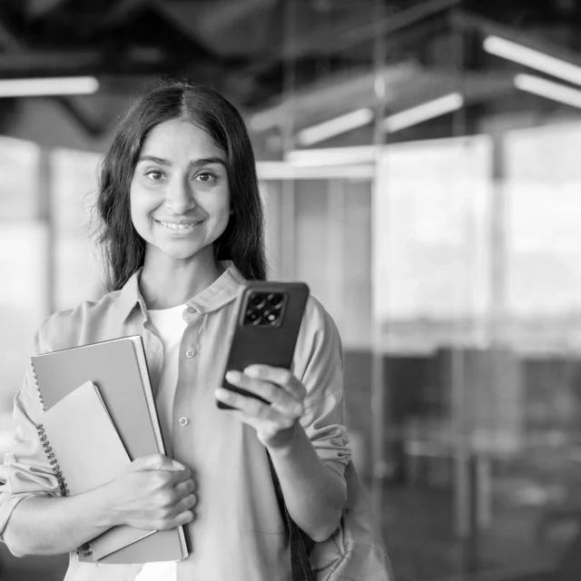 Student holding notebooks and a smartphone, smiling while standing in a modern learning environment.