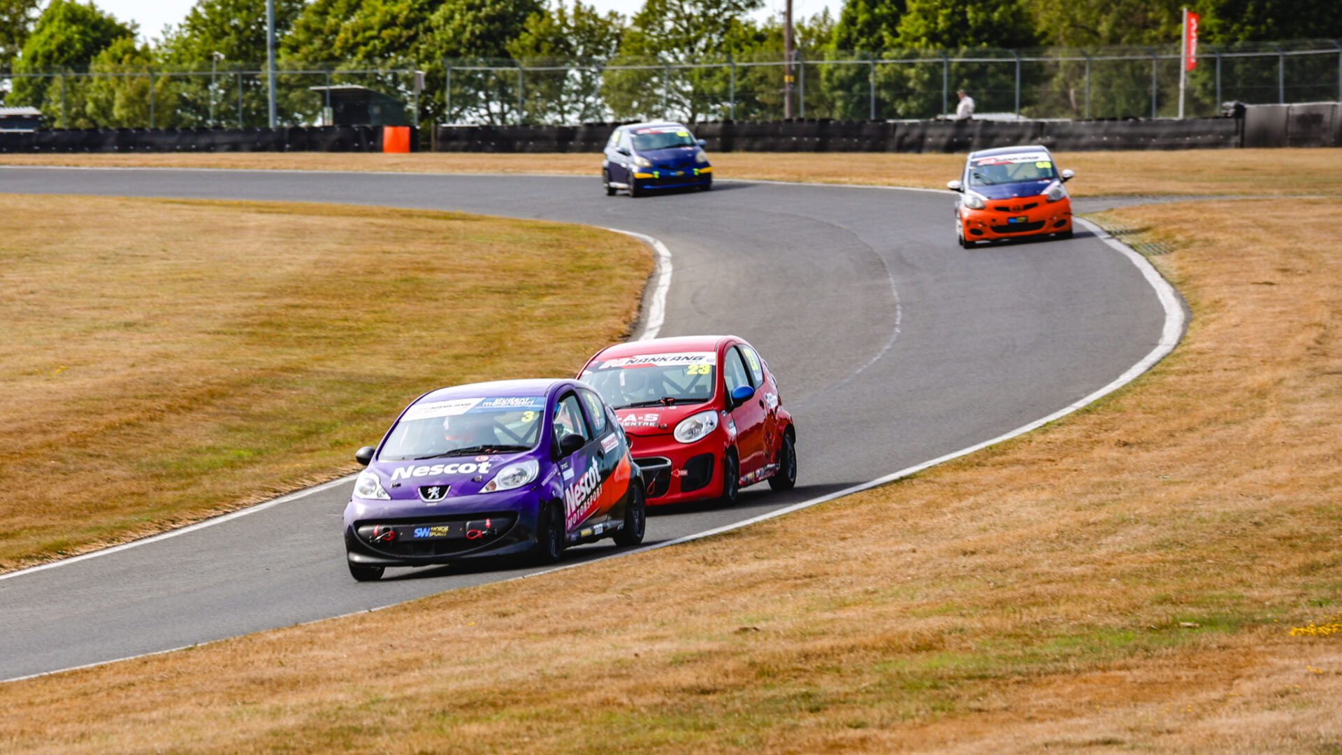 Nescot purple Peugeot race car leads a rival through a sweeping bend at Cadwell Park during a Student Motorsport race weekend, with additional competitors visible further along the track.