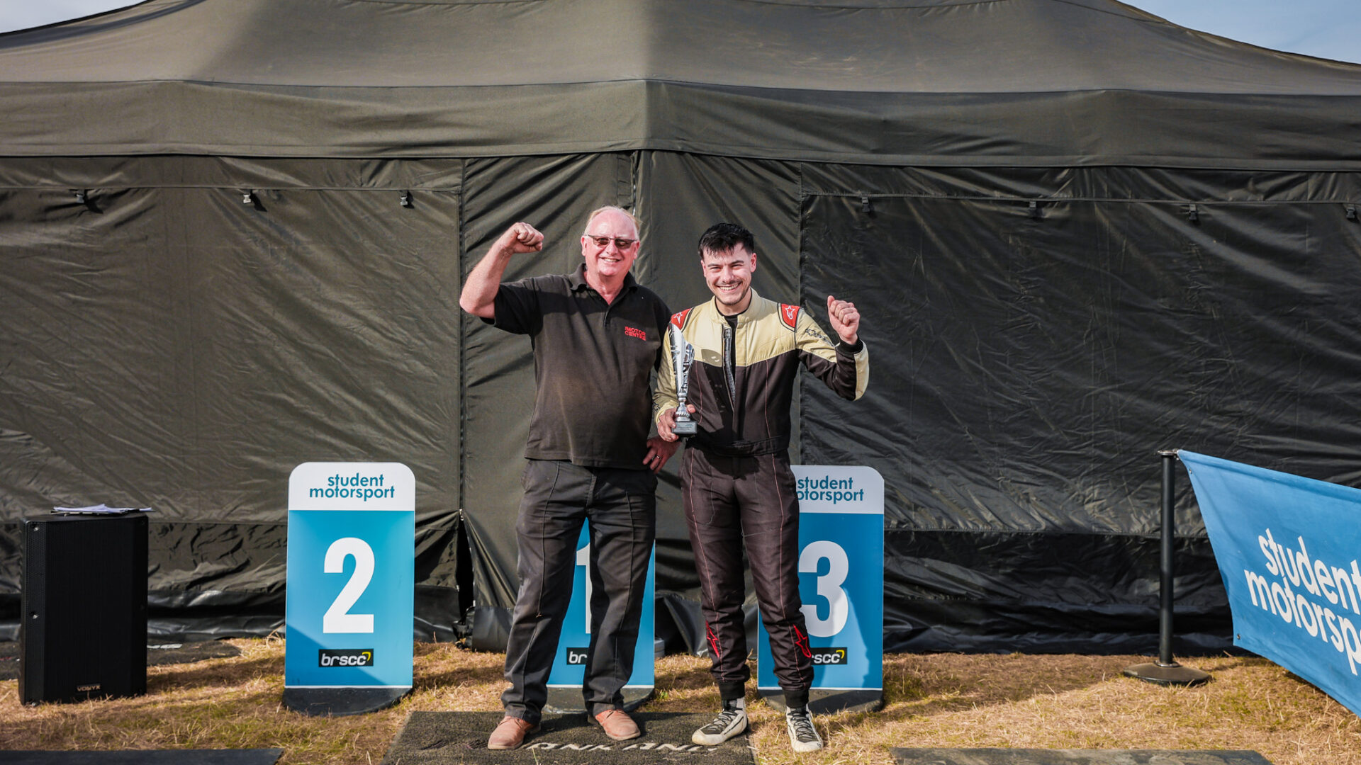 Nescot racing driver Elliot Lettis and Curriculum Manager George Keith celebrate a 3rd place podium at Cadwell Park as part of the Student Motorsport Competition. Both have one arm up in celebration with Elliot holding the trophy following a successful race weekend.