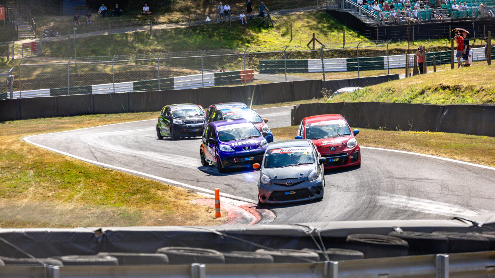 The purple Nescot Peugeot races through a bend at Cadwell Park alongside five other competitors during the Student Motorsport event, with trackside spectators and grandstands visible in the background.