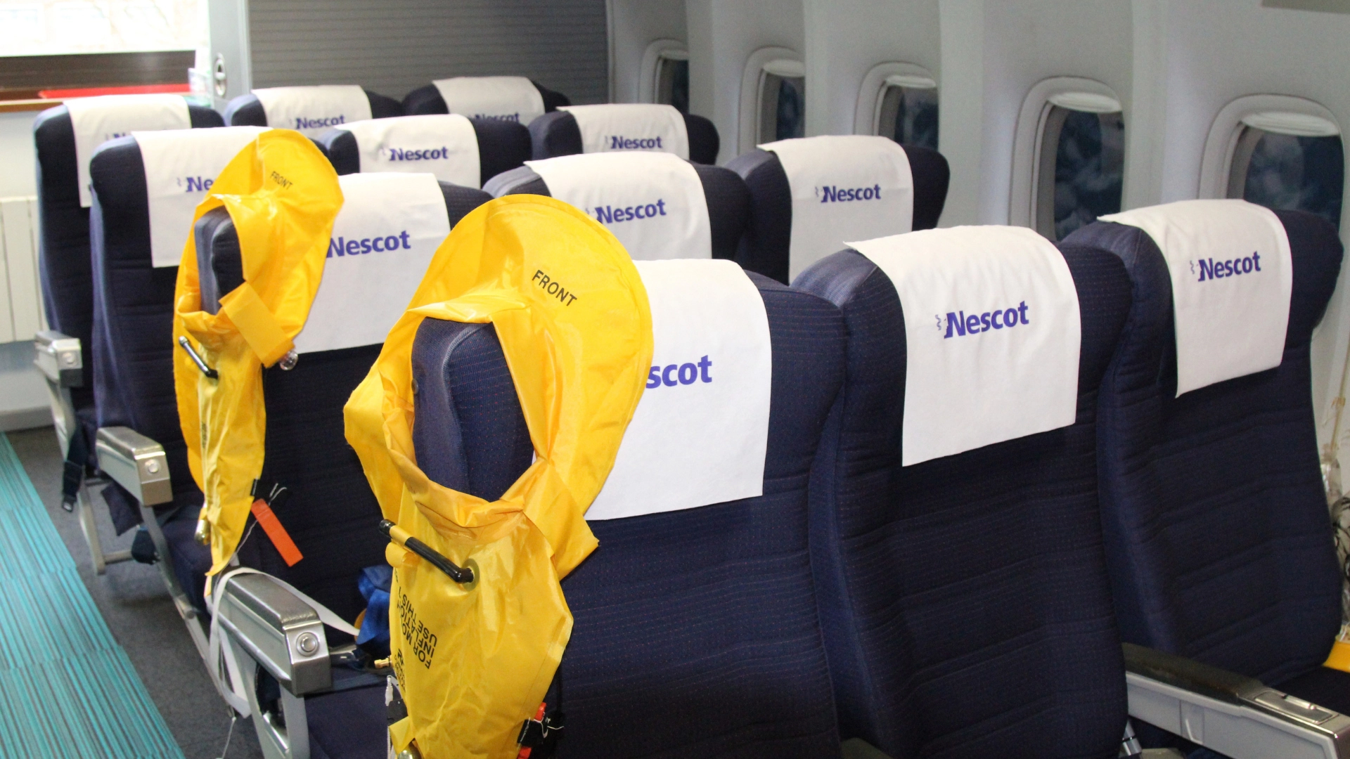 Airline cabin seats with the Nescot logo on the headrests. Lifejackets hanging on the end of the seats.