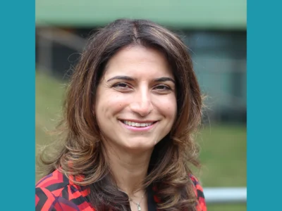 Head-and-shoulders portrait of Nescot's Head of Quality Improvement and Innovation with shoulder-length dark hair wearing a red and black geometric patterned blouse, photographed outdoors with a softly blurred green and grey background.