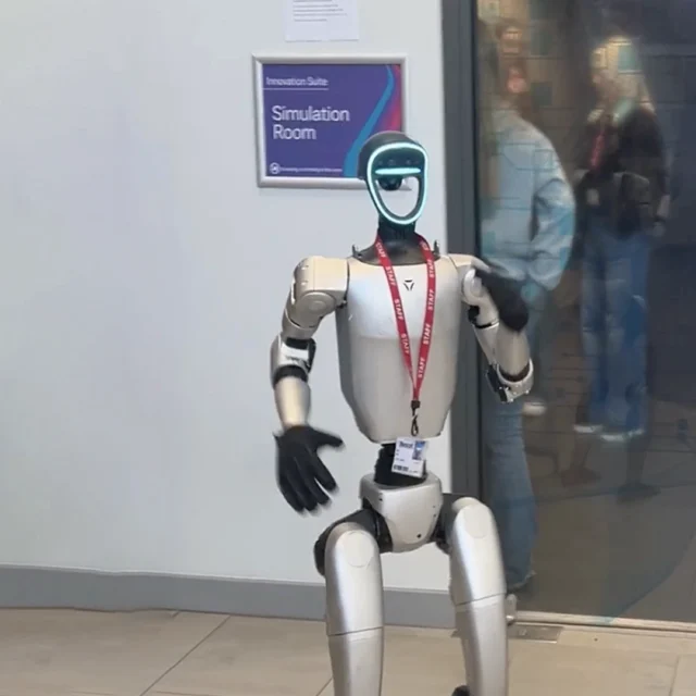 Humanoid robot standing outside a simulator room at Nescot with it's hands raised.
